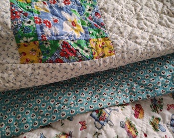 Op de afbeelding: Een doorgestikt stuk stof met een patchworkblok met bloemmotieven in rood, geel, groen en blauw. De quilt bevat een wit gedeelte met kleine schaarpictogrammen, een blauwgroene rand met witte bloemen en een witte basis met tuinmotieven.