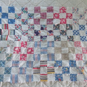 May include: A patchwork quilt featuring a grid of alternating white fabric squares and various patterned fabric squares in shades of blue, pink, red, yellow, and purple. The quilt is constructed from numerous small, square textile pieces.