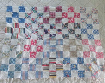 Op de afbeelding: Een patchwork quilt met een raster van afwisselende witte stoffen vierkanten en diverse stoffen vierkanten met patronen in tinten blauw, roze, rood, geel en paars. De quilt is opgebouwd uit talloze kleine, vierkante stukjes textiel.