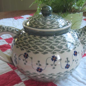 May include: A ceramic teapot featuring a cream-coloured base with a repeating dark green scale pattern on the top and bottom. The centre of the teapot is decorated with a horizontal band of small blue flowers with orange centres connected by thin green vines.