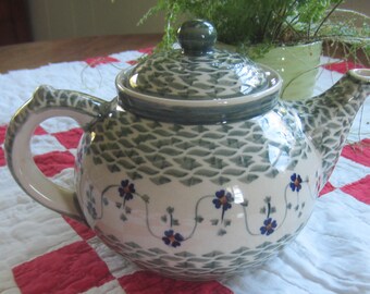 Op de afbeelding: Een keramische theepot met een crèmekleurige basis en een herhalend donkergroen schubpatroon aan de boven- en onderkant. Het midden van de theepot is versierd met een horizontale band van kleine blauwe bloemen met oranje harten verbonden door dunne groene ranken.