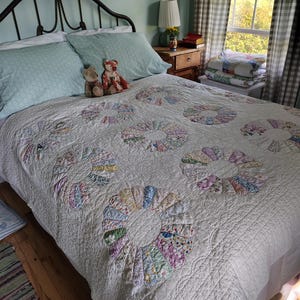 Puede incluir: Una colcha blanca con un patrón repetitivo de motivos circulares en forma de abanico, hechos de diversas telas con estampados florales en tonos pastel. La colcha tiene una superficie acolchada y texturizada sobre una cama con dos almohadas.