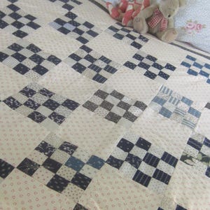 May include: A patchwork quilt featuring a repeating pattern of cream-colored fabric squares with small red floral prints alternating with dark blue and white checkered blocks. Two plush teddy bears sit on the quilt, one red and white, one tan.