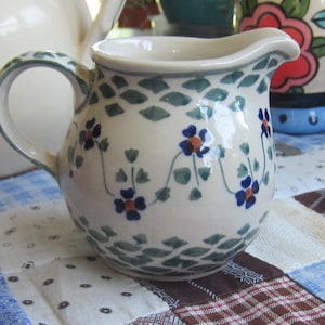 Op de afbeelding: Een keramieken roomkannetje met een crèmekleurige basis, voorzien van een handgeschilderd patroon van blauwe bloemen met oranje harten verbonden door groene ranken, en een decoratief groen schubbenmotief rond de bovenrand en de voet.