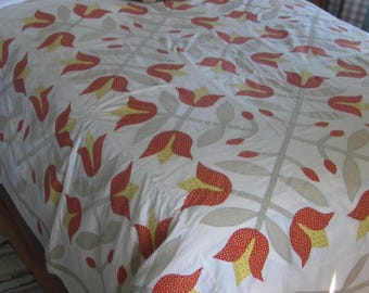 Op de afbeelding: Een witte bedsprei met een herhalend bloemenpatroon van oranje en gele tulpvormige motieven verbonden door lichtbeige stengels en bladeren. De sprei ligt over een bed met twee witte kussens en twee kleine knuffelberen.