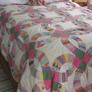 Peut inclure: Une courtepointe de style vintage avec un motif de doubles anneaux de mariage. La courtepointe présente un fond blanc avec des anneaux entrelacés roses, verts et en divers tissus à motifs. Deux ours en peluche sont sur les oreillers.
