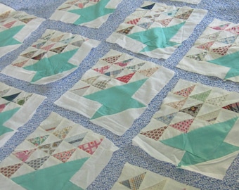 Op de afbeelding: Een quilt-top met een herhalend patroon van witte stoffen blokken met grote groenblauwe driehoeken en kleinere veelkleurige driehoeken met diverse prints. De blokken zijn geplaatst op een lichtblauwe stoffen achtergrond met een wit bloemenpatroon.