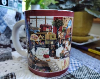 Op de afbeelding: Een keramische mok met een wit handvat en een donkerrode rand en voet. De buitenkant is voorzien van een gedetailleerde illustratie van een naaikamer, inclusief een vintage Singer-naaimachine, een rode naaidoos, garen en een kat.