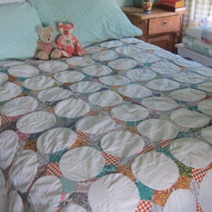 May include: A bed quilt featuring a repeating pattern of large white circular fabric patches stitched over a multicolored patchwork base. The quilt is spread across a bed with two light blue pillows and two small teddy bears sitting on top.