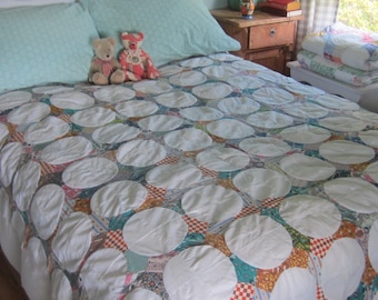 Op de afbeelding: Een bedsprei met een herhalend patroon van grote witte cirkelvormige stoffen lappen genaaid over een veelkleurige patchwork basis. De sprei ligt over een bed met twee lichtblauwe kussens en twee kleine teddyberen die erop zitten.