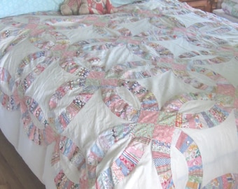 Op de afbeelding: Een quilt met een dubbel trouwringpatroon op een witte achtergrond, bestaande uit in elkaar grijpende ringen van diverse veelkleurige bloemrijke en bedrukte stofresten. Twee kleine knuffels liggen bovenop de doorgestikte sprei.