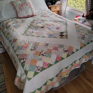 May include: A patchwork quilt featuring a central square of multi-colored fabric blocks surrounded by a wide white border and an outer edge of smaller, varied floral and geometric patterned squares in shades of pink, green, orange, and brown.