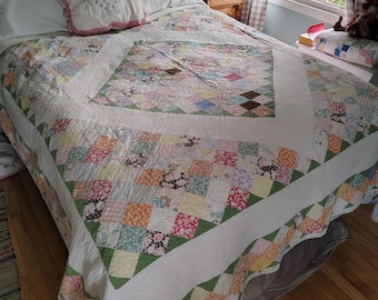 Op de afbeelding: Een patchwork sprei met een centraal vierkant van veelkleurige stoffen blokken, omringd door een brede witte rand en een buitenrand van kleinere, gevarieerde bloemrijke en geometrische vierkanten in tinten roze, groen, oranje en bruin.