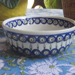 May include: A white ceramic bowl with a blue, red, and green circular pattern around the rim. The bowl is on a blue floral patterned tablecloth. A yellow pot and a wicker basket are in the background.