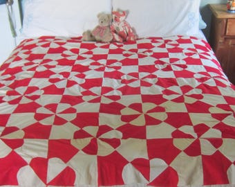 Op de afbeelding: Een quilt met een herhalend patroon van rode en witte stoffen blokken. Elk blok bevat gebogen vormen die een windmolen- of bloemmotief vormen. De quilt is plat uitgespreid en toont een consistente geometrische indeling over het gehele oppervlak.