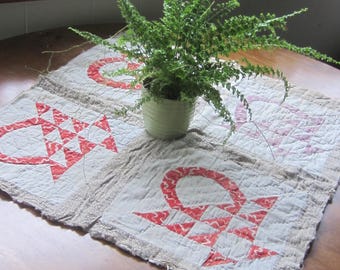Op de afbeelding: Een vierkante gewatteerde stoffen mat met vier afzonderlijke blokken met rood-witte mandpatronen, gescheiden door gestructureerde beige stoffen stroken. Een kleine groene varen in een pot staat in het midden van de mat met gerafelde randen.
