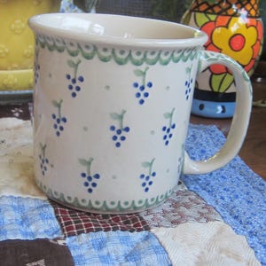 May include: A ceramic mug with a cream-coloured base featuring a hand-painted pattern of small blue grape clusters and green leaves. The rim and base are decorated with a scalloped green border, and the handle includes matching blue and green details.