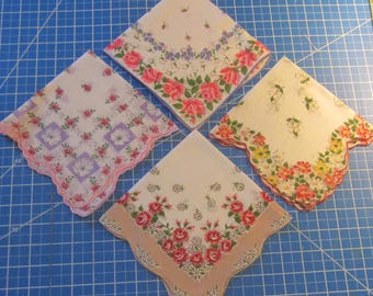 Op de afbeelding: Vier vierkante zakdoeken met bloemmotieven. Eén heeft roze rozen en blauwe bloemen, één heeft paarse vierkanten met witte bloemen, één heeft gele en oranje bloesems en één heeft rode rozen met een beige rand. Allemaal hebben ze geschulpte randen.