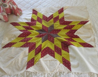 Op de afbeelding: Een vierkant quiltblok met een centraal achtpuntig sterpatroon bestaande uit ruitvormige stoffen stukjes in tinten bordeauxrood, geel en donkerpaars, tegen een effen gebroken witte achtergrond met een roze en witte teddybeer met patroon.