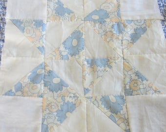 Op de afbeelding: Een vierkant stoffen quiltblok met een patchworkontwerp van effen witte vierkanten en driehoeken die afwisselen met een bloemenprintstof in tinten lichtblauw en bleekgeel. Het patroon bestaat uit aan elkaar genaaide geometrische vormen.