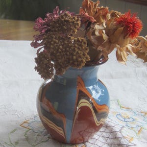 May include: A small, blue and brown ceramic vase with a rounded shape, containing dried flowers. The vase has a glossy finish with a swirled pattern of brown, orange, and cream. The flowers are various shades of brown, red, and tan.