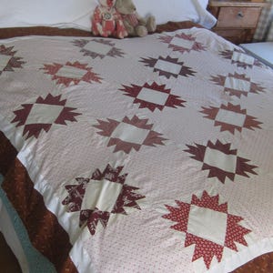 May include: A patchwork quilt featuring a repeating star pattern in shades of red, maroon, and cream. The quilt is constructed from various printed fabrics, including small floral and geometric motifs, with a solid brown border along the bottom edge.