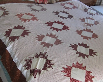 Op de afbeelding: Een patchwork quilt met een herhalend sterpatroon in tinten rood, bordeaux en crème. De quilt is gemaakt van diverse bedrukte stoffen, waaronder kleine bloemmotieven en geometrische patronen, met een bruine rand aan de onderzijde.