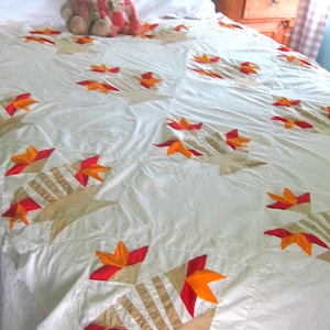 May include: Cream-colored quilt with a starburst pattern featuring red, orange, and tan accents. The quilt is on a bed with white sheets. Two teddy bears are sitting on the quilt.
