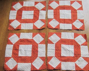 Op de afbeelding: Vier vierkante quiltblokken met een geometrisch patroon van rode stof met een kleine witte bloemenprint en effen witte stoffen vierkanten. Elk blok is opgebouwd met een centraal wit vierkant omringd door rode en witte geometrische vormen.
