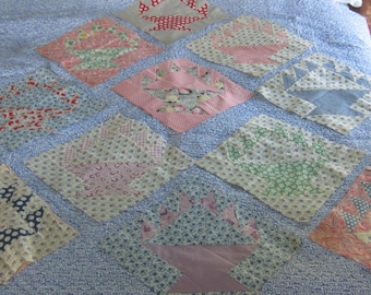 Op de afbeelding: Een quilt met een blauw-witte achtergrond met een patroon en meerdere vierkante stoffen blokken in een raster. Elk blok toont een samengesteld mandontwerp in verschillende kleuren, waaronder roze, rood, groen en blauw met bloemen- en geometrische prints.