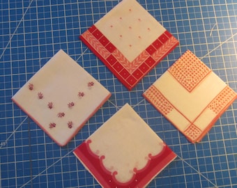 Op de afbeelding: Vier vierkante stoffen zakdoeken gerangschikt op een blauw rasteroppervlak. Elke doek heeft een eigen rood-wit patroon, waaronder bloemmotieven, geometrische randen, stippenontwerpen en geschulpte randen, allemaal in een ruitvorm geplaatst.