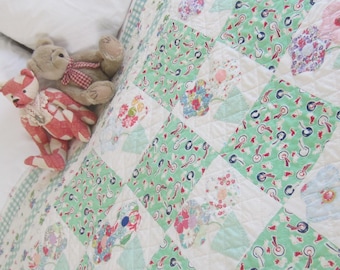 Op de afbeelding: Een patchwork quilt met een herhalend patroon van groene, witte en bloemen vierkanten. Twee teddyberen liggen op de quilt. Eén beer is bruin met een rood-wit geblokte strik, de andere is rood-wit geblokt.