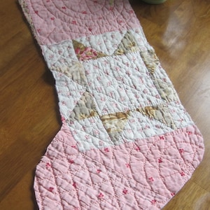 May include: A quilted Christmas stocking featuring a pink fabric with a small red floral print on the cuff and toe. The centre panel displays a white fabric with a star pattern in shades of beige, brown, and pink, all finished with detailed stitching.