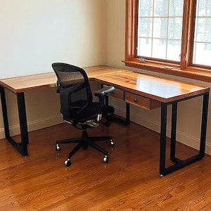 May include: L-shaped wooden desk with black metal legs and two drawers. A black mesh office chair sits in front of the desk. The desk is in a room with hardwood floors and a window.