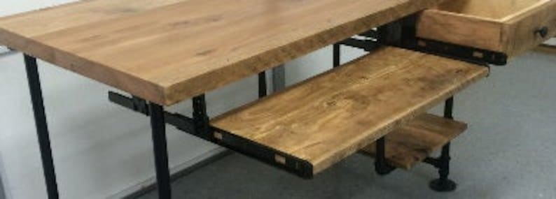 Op de afbeelding: Een houten bureau met een zwart metalen frame en een uittrekbare plank. Het bureau heeft een natuurlijke houten afwerking en een rustieke uitstraling.
