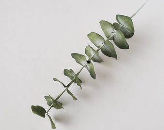 May include: A single artificial eucalyptus stem featuring a central wire branch with multiple rounded, sage green leaves arranged in an alternating pattern along the length of the stem, showcasing a matte finish and realistic leaf textures.