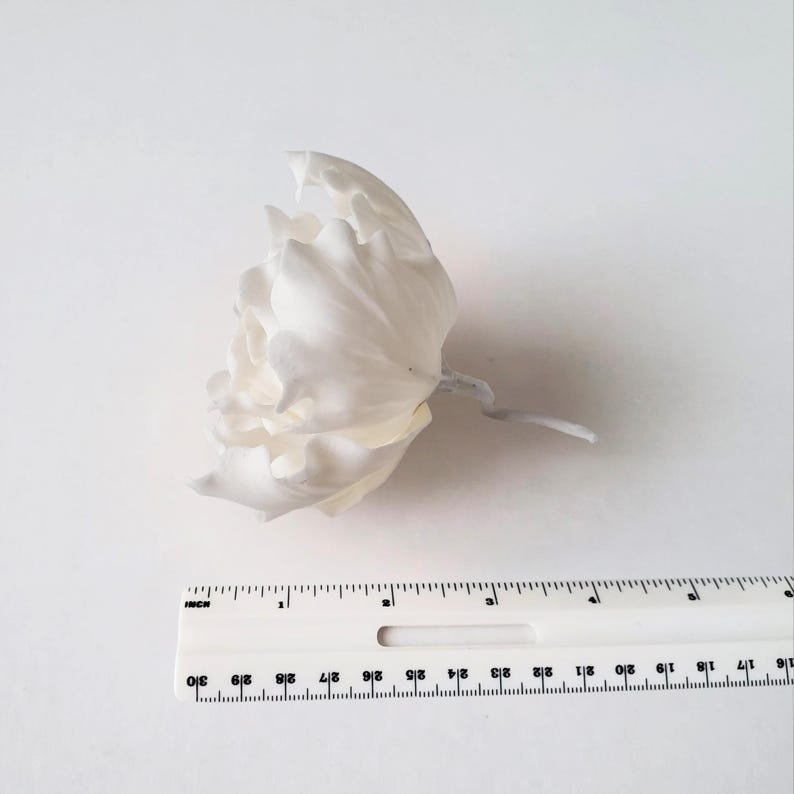 May include: A matte white artificial flower with layered, ruffled petals and a short stem, positioned above a white plastic ruler that shows the flower head measures approximately 3 inches in width from the outer edges of the petals.