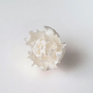 May include: A white flower ornament featuring multiple layers of delicate, ruffled petals. The structure is dense and compact, with a smooth, matte finish throughout. The petals curve outward from the center, creating a complex, layered appearance.