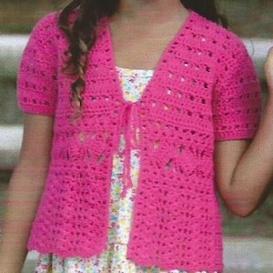 Puede incluir: Un cárdigan rosa brillante, de manga corta, tejido a ganchillo, con cierre de lazo. El diseño calado del suéter es visible. El cárdigan se lleva sobre un vestido con estampado floral.