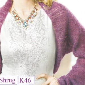May include: A purple knitted shrug with a delicate lace pattern. The shrug is worn over a white top with a beaded necklace.