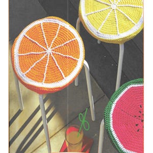 May include: Three crocheted stool covers in the shape of fruit slices. The covers are orange, yellow, and pink, and they are all crocheted with white yarn. The stool covers are on white metal stools. The image also includes a glass of orange juice with a green straw and the text "H166".