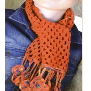 May include: A bright orange crochet scarf with a lacy pattern and large, colorful tassels. The scarf is worn around the neck of a person wearing a denim jacket. The text "CA98" is visible in the image.