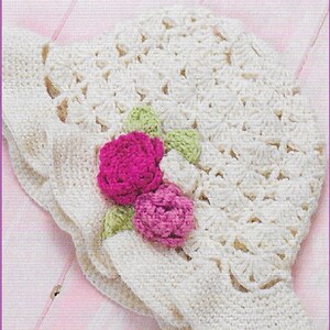 Puede incluir: Un sombrero blanco de ganchillo con un diseño floral. El sombrero tiene dos flores rosas y hojas verdes. El sombrero está sobre un fondo rosa. CC92