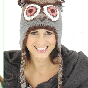 May include: A brown and grey crocheted owl hat with pink and white eyes. The hat has long, braided ties that hang down the sides.