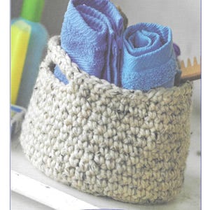 Op de afbeelding: Een beige gehaakte mand met een opgerolde blauwe handdoek erin. De mand heeft aan elke kant een handvat. De tekst "H8" is zichtbaar onder de mand.