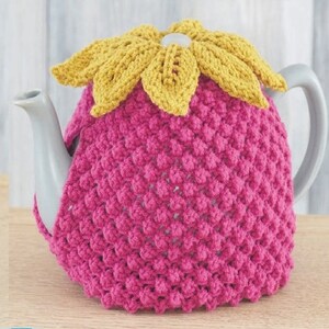 Op de afbeelding: Een roze gehaakte theepotwarmer met een gele gehaakte bloem erop. De warmer is ontworpen om te passen op een theepot van standaardformaat.