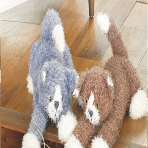 May include: Two knitted cat toys, one blue and one brown, with white fur accents. The cats are lying on a wooden surface and have a playful expression on their faces. The blue cat has a string tied around its paw.
