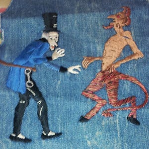 May include: Embroidered image on denim fabric depicting a man in a top hat and a devil. The man is wearing a blue jacket and black pants. The devil is red and has a tail.