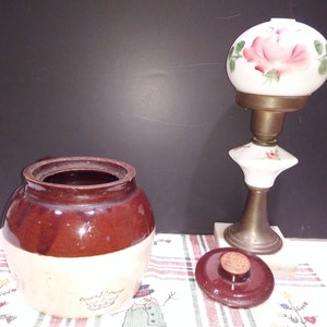 Puede incluir: Un tarro de cerámica esmaltada marrón y blanco con tapa. El tarro tiene un diseño floral en el lateral. La tapa es marrón y tiene un pomo en la parte superior. El tarro está sobre un mantel a rayas rojas y blancas. Una lámpara de vidrio blanco con un diseño de rosa rosa está en el fondo.