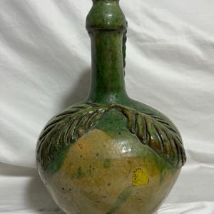 May include: A green and tan ceramic jug with a handle and a decorative leaf design. The jug has a rounded body and a long neck. The colors are earthy and the jug appears to be vintage.